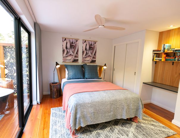 The bedroom. There is a queen sized bed with 2 pillows and 2 large cushions, There are 2 large pictures of Eucalyptus leaves on the wall above the bed. Beside the bed are bedside tables made of logs. Each has a bedside lamp on them. The flooring is wooden and there is a large grey rug under the bed. The bedroom has floor to ceiling curtains. There is a large glass sliding door which leads out to the back bath deck. There is an inbuilt desk with a bookshelf with books on above. There is a closet on the left side of the bed.