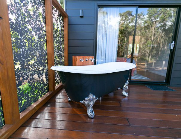 A large claw foot cast iron bath on the back deck of the chalet. There is a privacy screen on the left hand side. The path taps are mounted on a wooden box. The bath is located 1 metre from the sliding door to the bedroom.