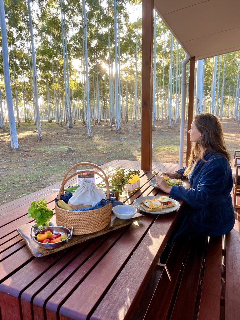 A woman in a blue bathrobe sitting on the front deck eating breakfast and looking at the view of the trees. There is a breakfast hamper on the table with a plate of cooked eggs on toast.