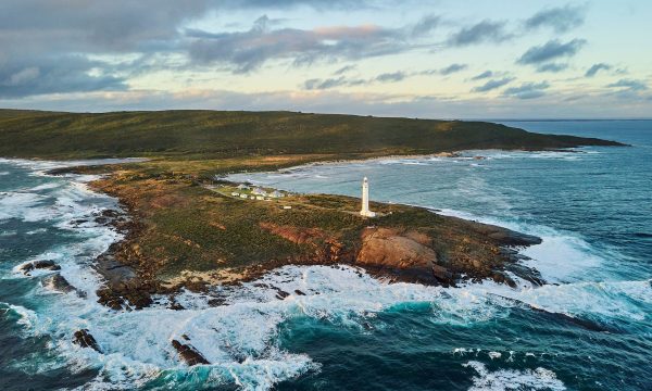 A drone image of Cape Leeuwin Lighthouse. The lighthouse is on the end of a piece of land that juts out into the ocean. The coastline is very rocky and the waves are breaking on the rocks.